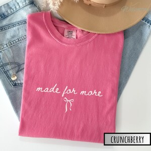 Puede incluir: Una camiseta rosa con la frase "made for more" en escritura blanca y un lazo. La camiseta est&aacute; doblada sobre unos vaqueros azules, con un sombrero beige y un collar dorado.