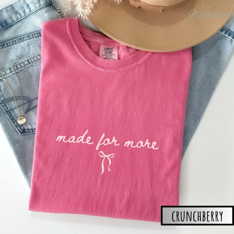 Puede incluir: Una camiseta rosa con la frase "made for more" en escritura blanca y un lazo. La camiseta est&aacute; doblada sobre unos vaqueros azules, con un sombrero beige y un collar dorado.