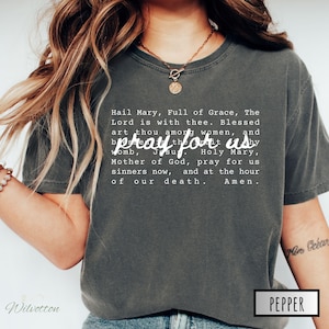 May include: Dark grey t-shirt with the text "Hail Mary, Full of Grace, The Lord is with thee. Blessed art thou among women, and pray for us by Jesu. Holy Mary, Mother of God, pray for us sinners now, and at the hour of our death. Amen."
