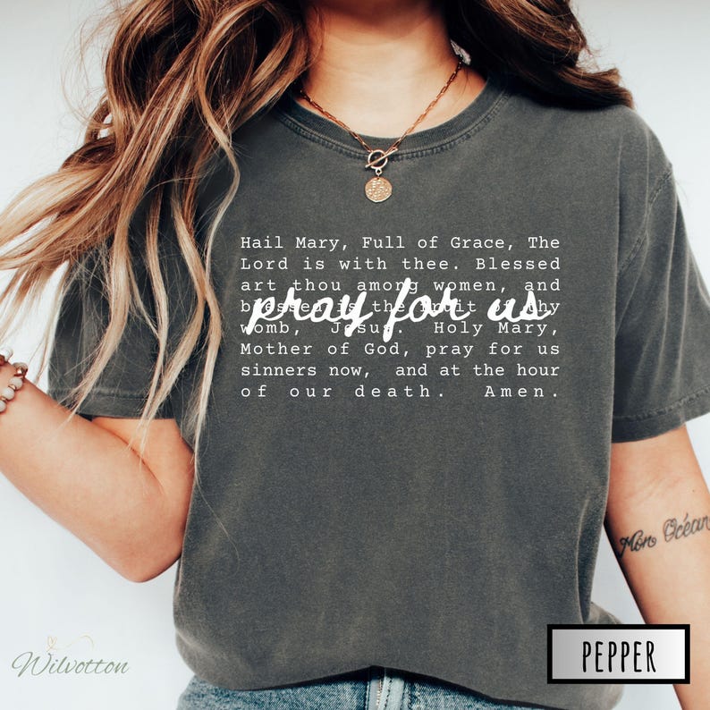 May include: Dark grey t-shirt with the text "Hail Mary, Full of Grace, The Lord is with thee. Blessed art thou among women, and pray for us by Jesu. Holy Mary, Mother of God, pray for us sinners now, and at the hour of our death. Amen."