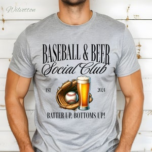 May include: A gray t-shirt with the text "BASEBALL & BEER Social Club" in black. The shirt also features a baseball glove with a baseball and a glass of beer. The text "EST" and "2024" are on either side of the glove and beer. The text "BATTER UP, BOTTOMS UP!" is at the bottom of the shirt.