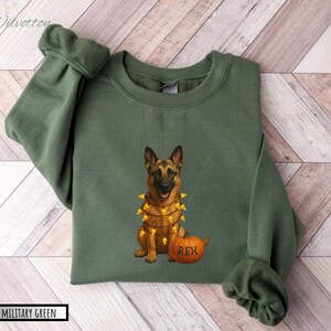 May include: Military green sweatshirt featuring a German Shepherd dog with a string of lights around its neck and a pumpkin with the name "Rex". The sweatshirt has long sleeves and a crew neck. The text "MILITARY GREEN" is at the bottom.