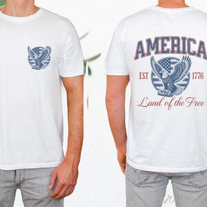 Puede incluir: Camiseta blanca con un gr&aacute;fico de &aacute;guila azul y rojo en la parte delantera y el texto "AMERICA EST 1776 Land of the Free" en la parte posterior.