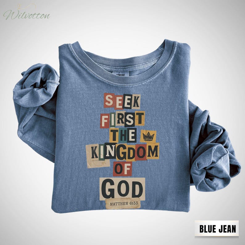 May include: Blue jean long-sleeve shirt featuring the text "SEEK FIRST THE KINGDOM OF GOD" in a colourful, cut-out letter design. The shirt also has the text "MATTHEW 6:33". The shirt is folded with rolled-up sleeves.
