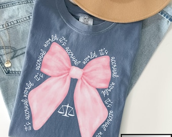 May include: A blue t-shirt with a pink bow and the text "it's accrual world it's accrual world it's accrual world it's accrual world" printed on it.
