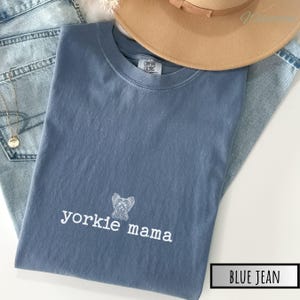 May include: A blue jean colored t-shirt with the words "yorkie mama" and a yorkshire terrier graphic in white. The shirt is folded and placed on top of a pair of blue jeans. A tan hat is partially visible in the background.