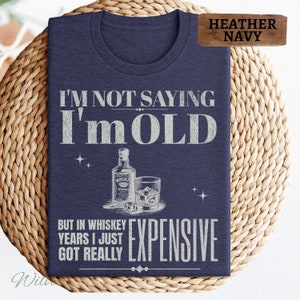 Puede incluir: Una camiseta azul marino jaspeada con un dise&ntilde;o gr&aacute;fico blanco. El gr&aacute;fico presenta una botella de whisky, un vaso de whisky con hielo y el texto "No estoy diciendo que soy viejo, pero en a&ntilde;os de whisky me he vuelto realmente caro."