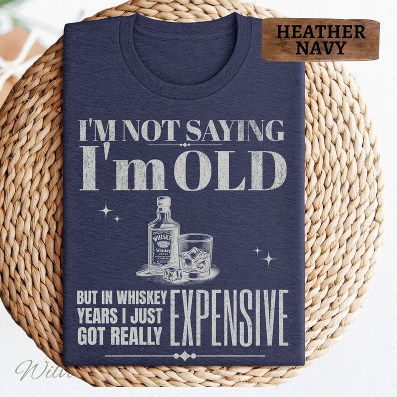 Puede incluir: Una camiseta azul marino jaspeada con un dise&ntilde;o gr&aacute;fico blanco. El gr&aacute;fico presenta una botella de whisky, un vaso de whisky con hielo y el texto "No estoy diciendo que soy viejo, pero en a&ntilde;os de whisky me he vuelto realmente caro."