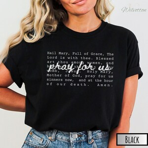 May include: Black t-shirt with the text "pray for us" in a stylized font. The shirt also features the Hail Mary prayer in white text. The shirt is a crew neck and is made of a soft material.