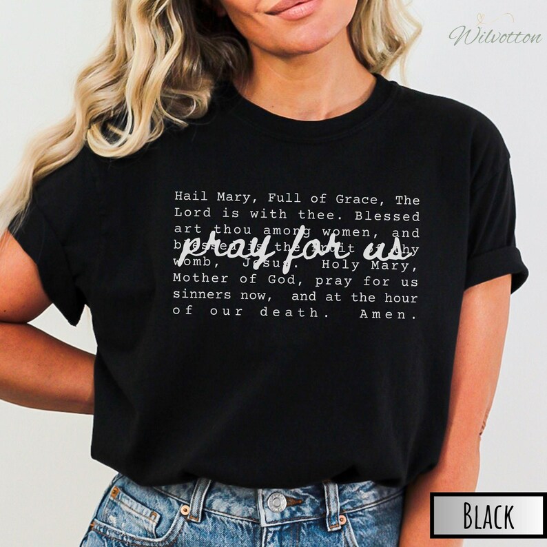 May include: Black t-shirt with the text "pray for us" in a stylized font. The shirt also features the Hail Mary prayer in white text. The shirt is a crew neck and is made of a soft material.