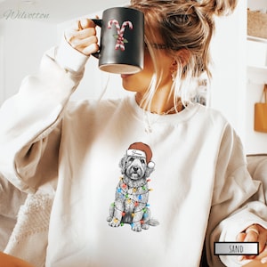 May include: Sand-colored sweatshirt featuring a graphic of a dog wearing a Santa hat and Christmas lights. The dog's name, "Bruno", is printed on the hat. A black mug with candy cane and holly berry design is held up. The word "Sand" is printed on the bottom right.
