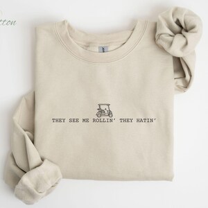 Pu&ograve; includere: Una felpa beige con una grafica nera di un golf cart e il testo "They see me rollin' they hatin'".