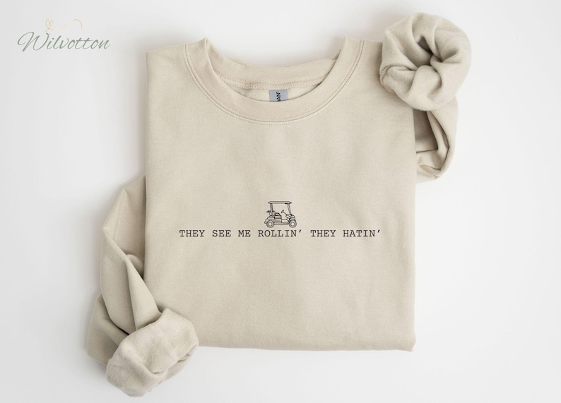 Pu&ograve; includere: Una felpa beige con una grafica nera di un golf cart e il testo "They see me rollin' they hatin'".