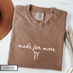 Puede incluir: Camiseta Comfort Colors color espresso con la frase "made for more" en escritura blanca y un gr&aacute;fico de lazo. La camiseta est&aacute; doblada y tiene cuello redondo. La etiqueta dice S-P-CH.