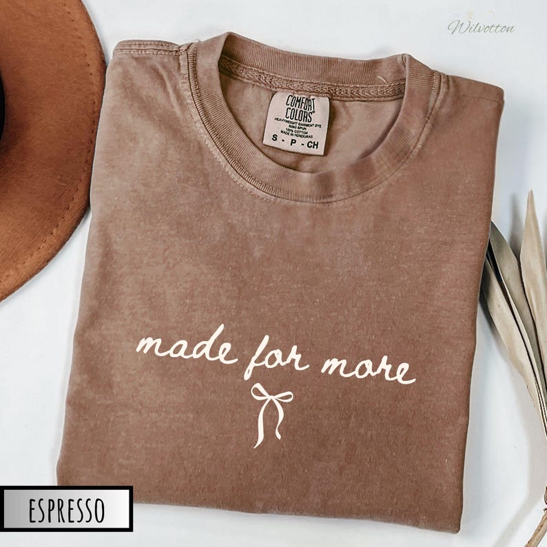 Puede incluir: Camiseta Comfort Colors color espresso con la frase "made for more" en escritura blanca y un gr&aacute;fico de lazo. La camiseta est&aacute; doblada y tiene cuello redondo. La etiqueta dice S-P-CH.