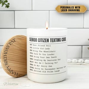 May include: A white candle with a lit wick, featuring the text "SENIOR CITIZEN TEXTING CODE." A wooden lid with "HAPPY BIRTHDAY PAT" is next to the candle. The candle is on a white surface.
