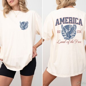 Puede incluir: Una camiseta blanca de gran tama&ntilde;o con un gr&aacute;fico de &aacute;guila azul y rojo en la espalda. El gr&aacute;fico incluye el texto "AMERICA EST 1776 Land of the Free". La parte delantera de la camiseta tiene una versi&oacute;n m&aacute;s peque&ntilde;a del gr&aacute;fico del &aacute;guila.