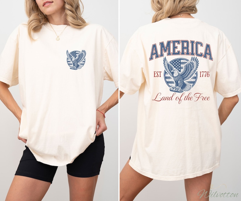 Puede incluir: Una camiseta blanca de gran tama&ntilde;o con un gr&aacute;fico de &aacute;guila azul y rojo en la espalda. El gr&aacute;fico incluye el texto "AMERICA EST 1776 Land of the Free". La parte delantera de la camiseta tiene una versi&oacute;n m&aacute;s peque&ntilde;a del gr&aacute;fico del &aacute;guila.