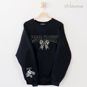 May include: Black sweatshirt with the text "MOTO NANNY" in white block letters and a checkered bow graphic. The sleeve features a motorcycle graphic with the name "Jaxson" and the number "45" in white.