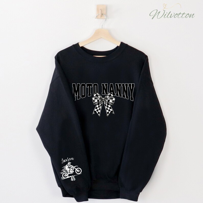 May include: Black sweatshirt with the text "MOTO NANNY" in white block letters and a checkered bow graphic. The sleeve features a motorcycle graphic with the name "Jaxson" and the number "45" in white.
