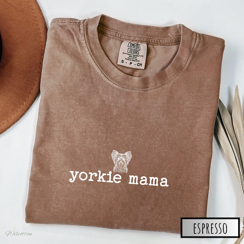 May include: Espresso-colored t-shirt with the text "yorkie mama" and a Yorkshire Terrier graphic. The shirt is a Comfort Colors brand, size small. The shirt is folded and displayed on a white surface.