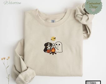 May include: Sand-colored sweatshirt featuring an embroidered Halloween design. The design includes a black and brown dog, a ghost, a campfire, a full moon, and a jack-o'-lantern. The sweatshirt has a crew neck and long sleeves. The word "SAND" is printed on the bottom right.