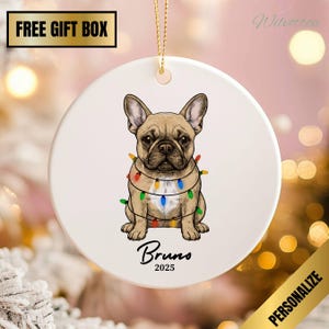 May include: A round, white ceramic ornament featuring a tan French Bulldog with a string of colorful Christmas lights. The dog's name, "Bruno," and the year "2025" are written below. A gold cord is attached for hanging.