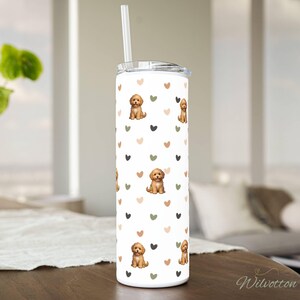 May include: A white tumbler with a repeating pattern of brown puppies and hearts in shades of brown, green, and black. The tumbler has a silver lid and a clear straw. The tumbler is sitting on a wooden surface.
