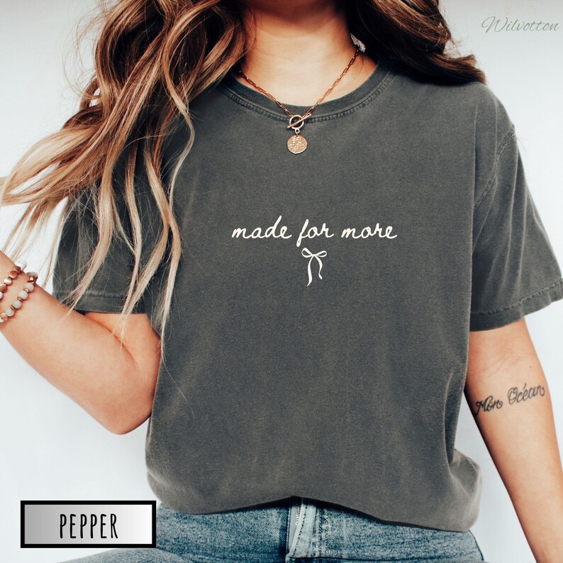 Puede incluir: Camiseta gris oscuro con la frase "made for more" en escritura blanca y un lazo gr&aacute;fico. La palabra "Pepper" est&aacute; en un cuadro blanco en la parte inferior. La camiseta tiene mangas cortas y cuello redondo.