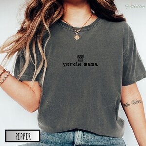 May include: A dark gray t-shirt with the text "yorkie mama" and a small dog graphic. The shirt is a relaxed fit and has short sleeves. The word "pepper" is in a white box at the bottom of the image.