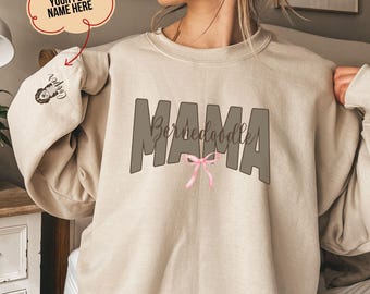May include: Beige sweatshirt with the text "MAMA Bernedoodle" in brown letters with a pink bow in the middle. The sweatshirt has a pocket on the left sleeve with the text "Cooper" in black.