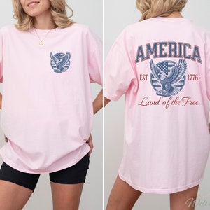 Puede incluir: Camiseta rosa de gran tama&ntilde;o con un gr&aacute;fico de &aacute;guila azul y rojo en la espalda. El gr&aacute;fico incluye el texto "AMERICA EST 1776 Land of the Free".