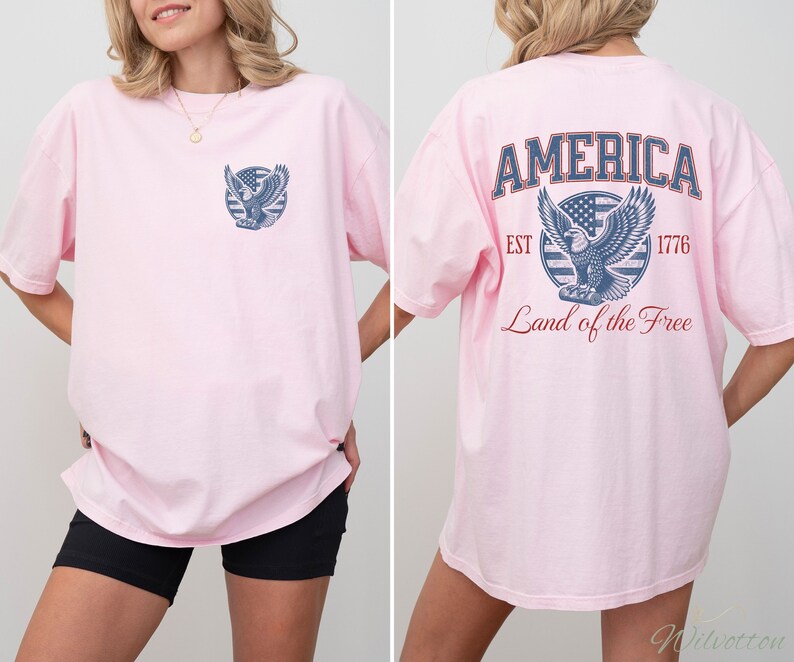 Puede incluir: Camiseta rosa de gran tama&ntilde;o con un gr&aacute;fico de &aacute;guila azul y rojo en la espalda. El gr&aacute;fico incluye el texto "AMERICA EST 1776 Land of the Free".