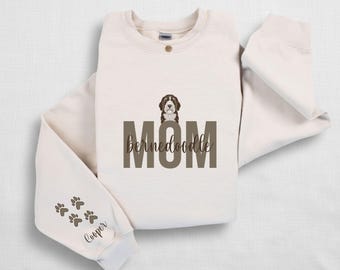 May include: Sand-colored sweatshirt with a brown and white Bernedoodle graphic above the word "MOM" in brown block letters. The word "bernedoodle" is below "MOM". The left sleeve has paw prints and the name "Cooper".
