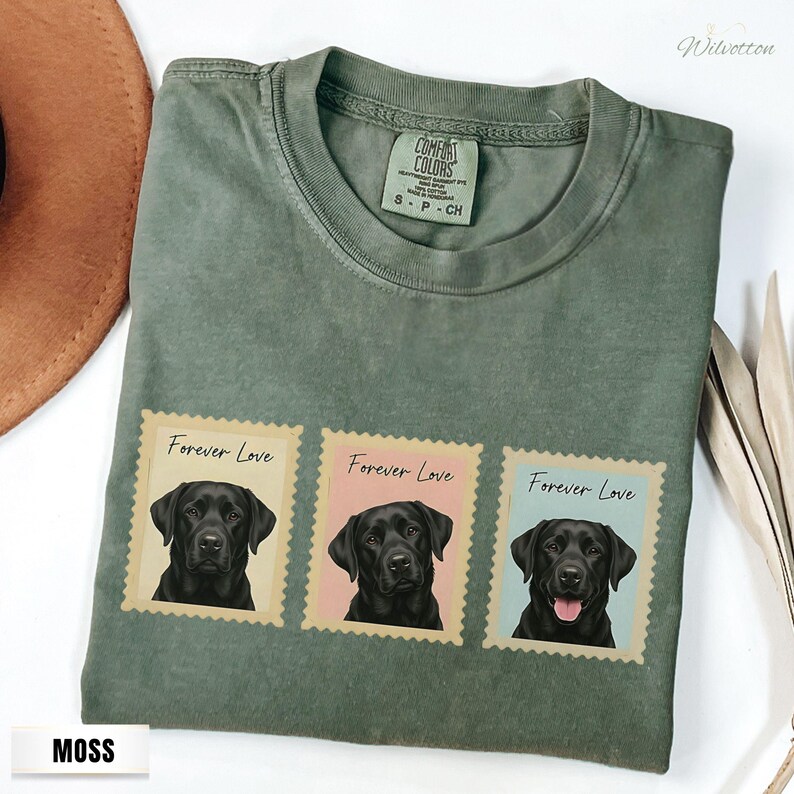 May include: Moss green t-shirt with three postage stamp designs featuring black Labrador Retrievers. Each stamp has the words "Forever Love" printed above the dog's portrait. The shirt is made of a soft, comfortable material.