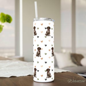 May include: A white tumbler with a lid and straw, featuring a repeating pattern of brown dogs and hearts in various colors. The tumbler is cylindrical and sits on a wooden surface.