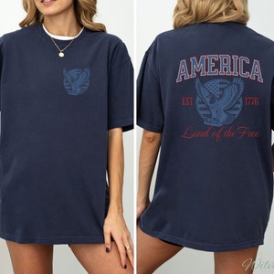 Puede incluir: Una camiseta azul marino con un gr&aacute;fico de un &aacute;guila y las palabras "AMERICA EST 1776 Land of the Free" impresas en la espalda.