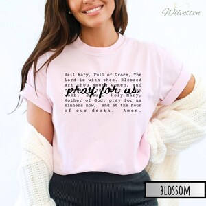 May include: Light pink t-shirt with the text "Hail Mary, Full of Grace, The Lord is with thee. Blessed art thou among women, and pray for us Holy Mary, Mother of God, pray for us sinners now, and at the hour of our death. Amen."