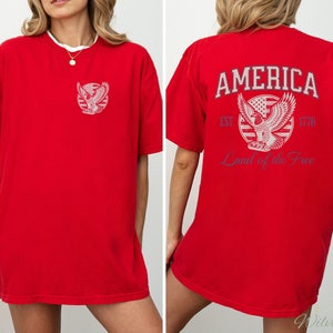 Puede incluir: Camiseta roja de gran tama&ntilde;o con un gr&aacute;fico de un &aacute;guila y el texto "AMERICA EST. 1776 Land of the Free" en la parte posterior.