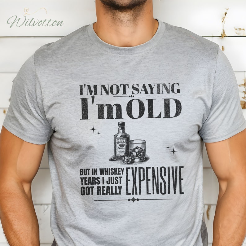 Puede incluir: Camiseta gris con un dicho humor&iacute;stico sobre el envejecimiento, "No estoy diciendo que soy viejo, pero en a&ntilde;os de whisky me he vuelto realmente caro." La camiseta presenta una ilustraci&oacute;n en blanco y negro de una botella de whisky y un vaso con cubitos de hielo.