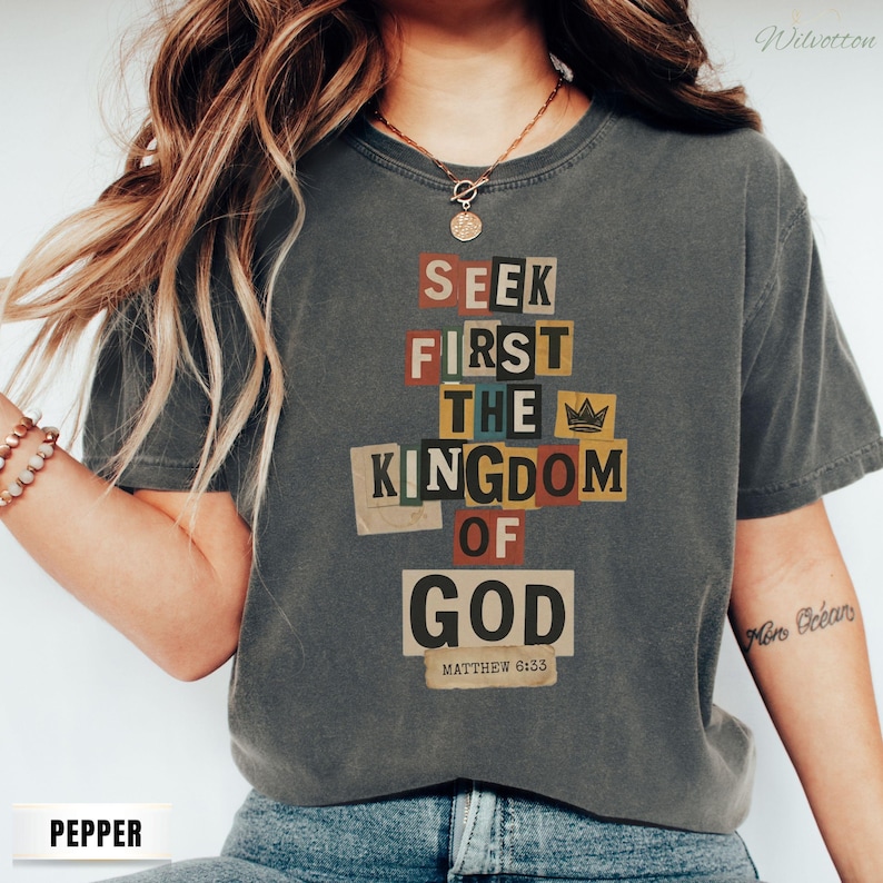 May include: A dark grey t-shirt featuring the text "SEEK FIRST THE KINGDOM OF GOD" in a collage style. The text is in various colours and fonts. The shirt also has the text "MATTHEW 6:33" at the bottom.