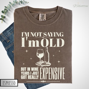 May include: Espresso-colored t-shirt with the text "I'M NOT SAYING I'm OLD BUT IN WINE YEARS I JUST GOT REALLY EXPENSIVE." The shirt features a wine glass, bottle, and food graphic. The shirt is folded on top of blue jeans.