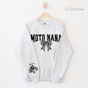 May include: A light gray sweatshirt with the text "MOTO NANA" in black block letters, a checkered bow, and a motorcycle graphic with the name "Jaxson" and the number "45". The sweatshirt is hanging on a wooden hanger.
