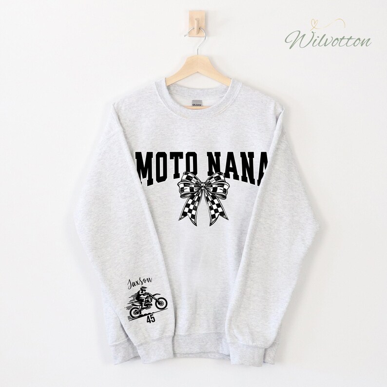 May include: A light gray sweatshirt with the text "MOTO NANA" in black block letters, a checkered bow, and a motorcycle graphic with the name "Jaxson" and the number "45". The sweatshirt is hanging on a wooden hanger.