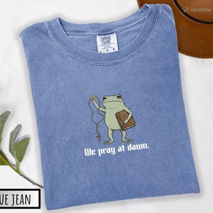 May include: A blue jean colored t-shirt with a graphic of a green frog holding a brown book and a rosary. Below the frog, the text reads "We pray at dawn." The shirt features a Comfort Colors brand label at the neck indicating a size large.