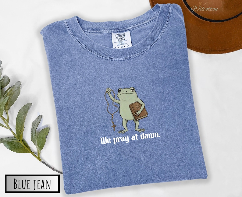 May include: A blue jean colored t-shirt with a graphic of a green frog holding a brown book and a rosary. Below the frog, the text reads "We pray at dawn." The shirt features a Comfort Colors brand label at the neck indicating a size large.