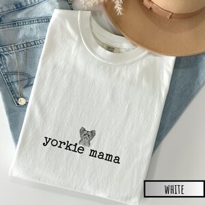 May include: White t-shirt with the text "yorkie mama" and a graphic of a Yorkshire Terrier. The shirt is folded on top of blue jeans, with a gold necklace and a tan hat in the background.