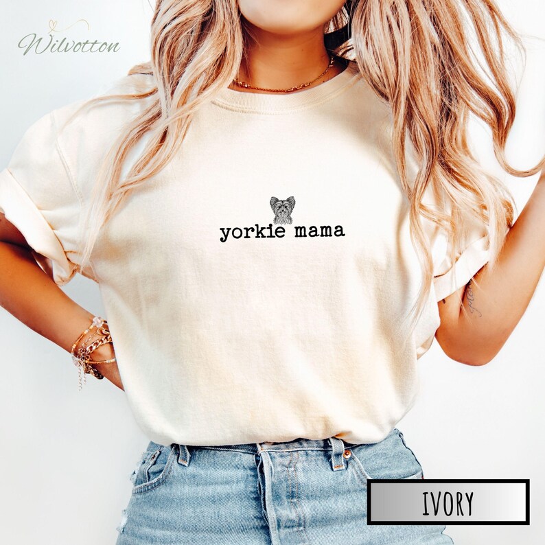 May include: Ivory-colored t-shirt with the text "yorkie mama" and a small illustration of a Yorkshire Terrier. The shirt is a light cream color and has rolled-up sleeves. The word "IVORY" is in a white box at the bottom.