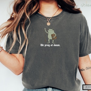 May include: A charcoal gray short-sleeve t-shirt featuring a graphic of a green frog holding a brown book. The frog is depicted with one arm raised. Below the illustration, the text reads "We pray at dawn." in a white, stylized serif font.