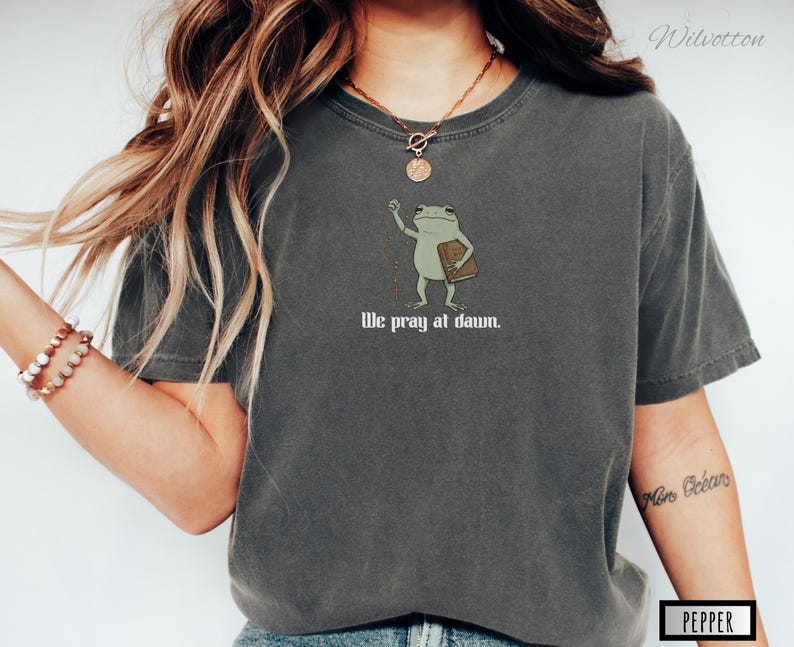 May include: A charcoal gray short-sleeve t-shirt featuring a graphic of a green frog holding a brown book. The frog is depicted with one arm raised. Below the illustration, the text reads "We pray at dawn." in a white, stylized serif font.